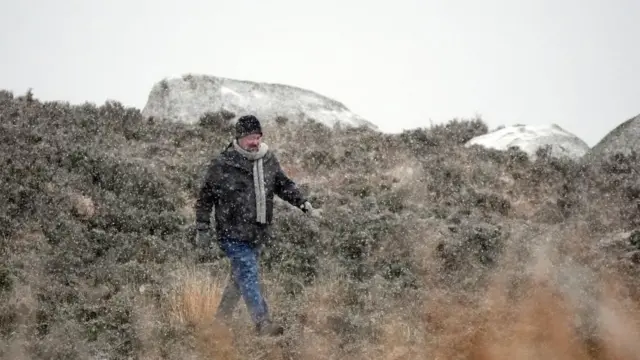 People are out walking at the Wicklow Gap mountain pass. There is snow in the foreground and some accumulation on the ground. A man is walking through a brushy area of ground wearing winter clothing.