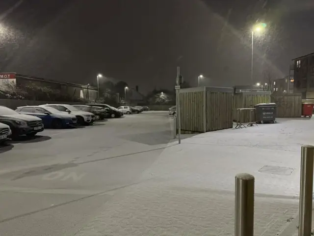 Snow covers a residential estate, where rows of cars are parked behind houses.