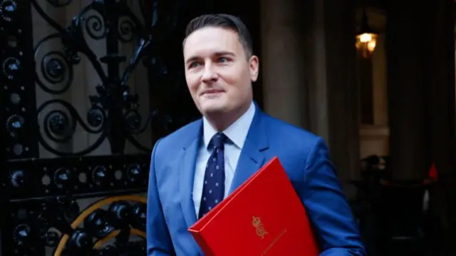 Wes Streeting walking out of a building while carrying a red government folder under his left arm. He's wearing a blue suit with a white shirt a dark blue, patterned tie. To his right, in the background, is a black iron gate