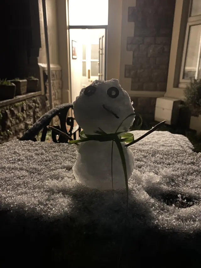 A tiny snowman with eyes made out of olives and a green scarf sits on a wall.