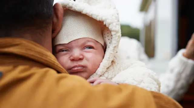 Baby in white winter wear being held by a man in yellow jacket with short brown hair, blurred background, with a feminine left arm leaning against a wall visible to the right of the frame