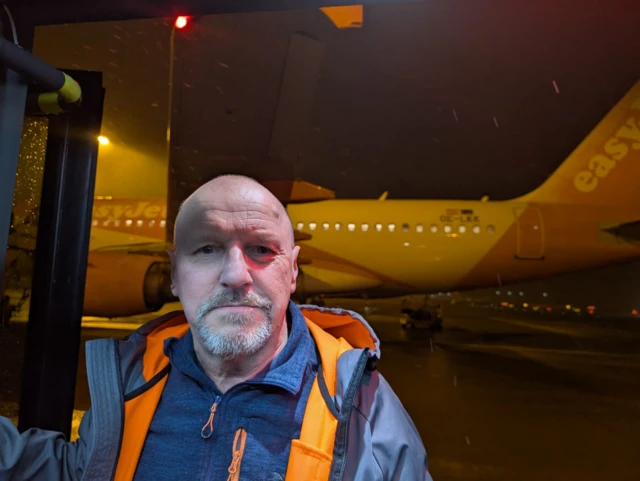 A man wearing a navy fleece and orange lined jacket poses for a selfie on a bus parked up near an easyjet plane.