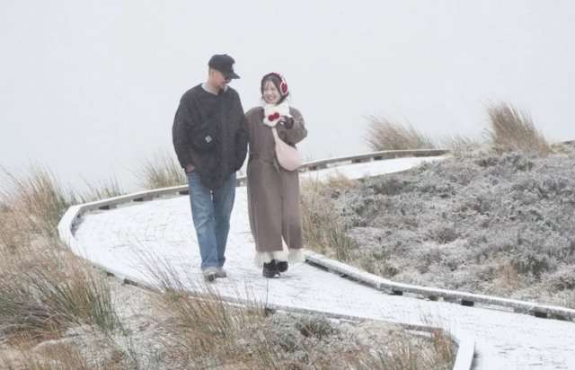 A couple in warm clothes hold arms as they walk a snow-covered mountain path in Wicklow, Ireland