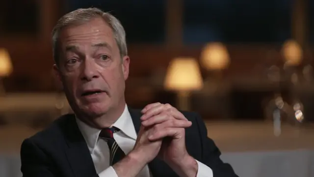 Nigel Farage wearing a suit and clasping his hands as he speaks to Laura Kuenssberg, Behind him, blurred, are tables covered in white cloth and lamps