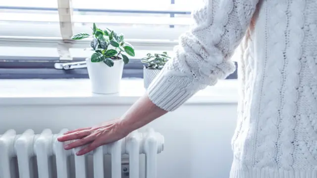 Woman in chunky white jumper holds her left hand over a white indoor heater while standing in front of a window. There are two plant pots on the windowsill