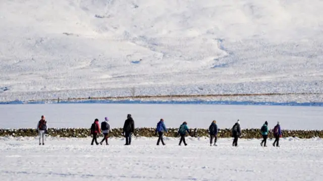 People walk through snow in the Pentland Hills, Balerno, Edinburgh. A group of 10 people, both male and female, walk through a snowy field with a snow-covered hill in the background