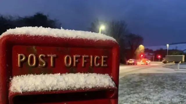 Snow covered red letter box in foreground to the left to the middle of the frame. To the right, in the background (blurred), cars at a red light driving over snowy road