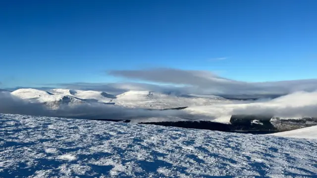 View of mountain tops covered in snow taken from the peak of A' Mharconaich. Sky is clear with only a few low clouds near the top of the peaks