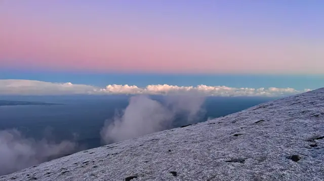 Frosty grass on a mountaintop is shown in this photo from Northern Ireland. There is a sunset with pink and blue sky in the background and clouds are seen at the edge of the mountain and further in the distance.