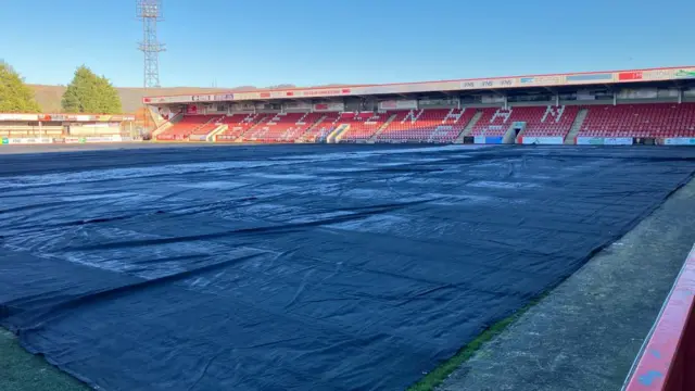 Cheltenham Town football pitch with stands in the background, large black ground sheet covers the grass with frost visible