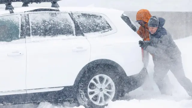 Two men in winter gear try to move a white car stuck in heavy snow