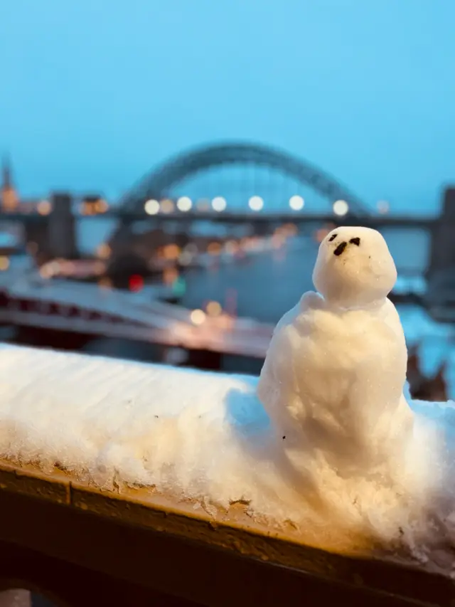 A very small snowman made on the handrail of a bridge, with the Tyne Bridge in the background