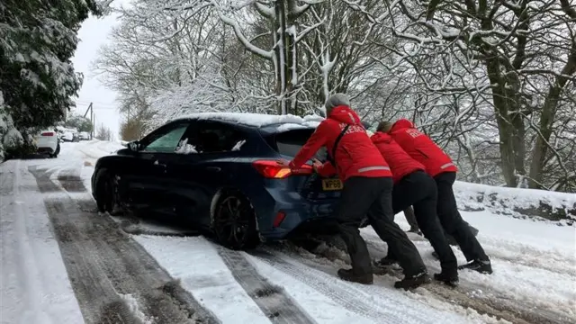 Three men in red jackets push a blue car along a snowy road.