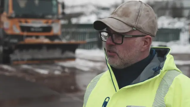 Mick Dand stands in front of a gritter. He wears a faded brown cap and high vis jacket.