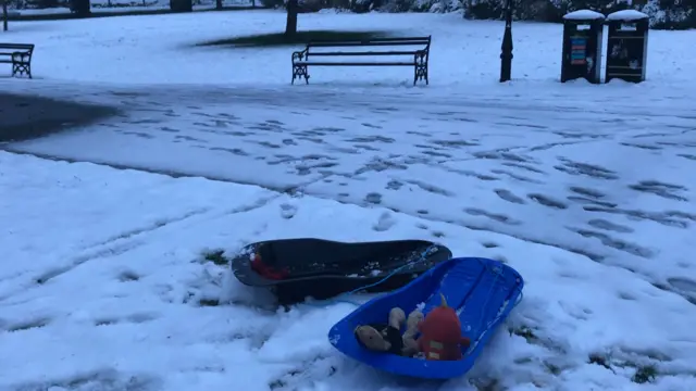 A blue and a black sledge side by side in a snowy park. The blue sledge has two children's teddies in it. Footsteps are trodden into the snow and a bench and bins sit beyond. the skies are still brightening and lights are on in a building behind trees.