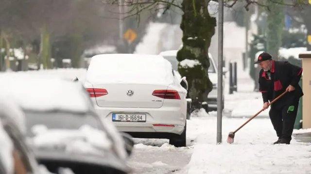 A man sweeps snow from a footpath next to cars parked on the road