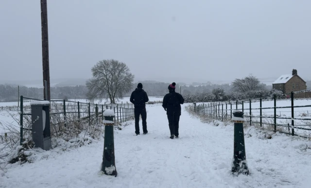 Two walkers wearing dark clothing on a snow-covered footpath, they are facing away from the camera and there are two bollards in the foreground