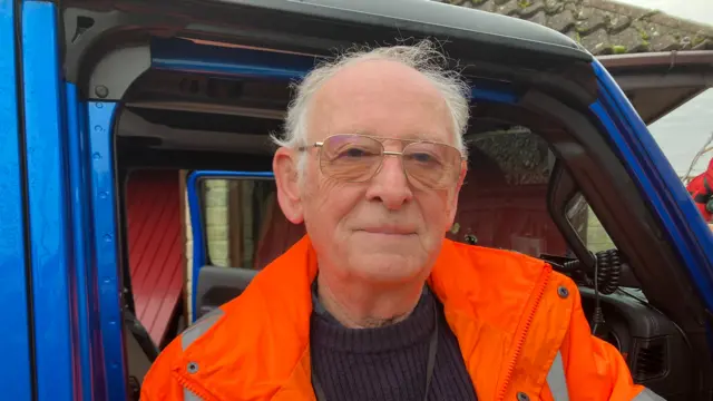 Jerry, pictured standing in the open door of his blue 4x4. He wears a bright orange high-vis jacket and large square glasses, and is looking directly at the camera.