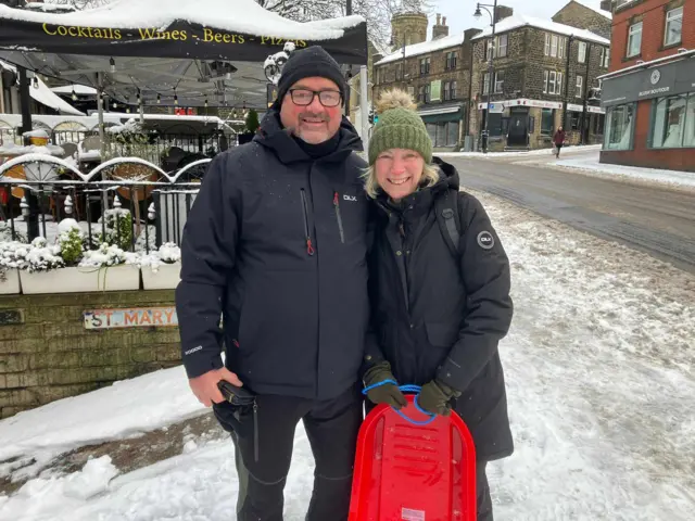 Paul and Nicola Parker smile together while Nicola holds a red sledge. The pair are stood on a snow-covered roadside.