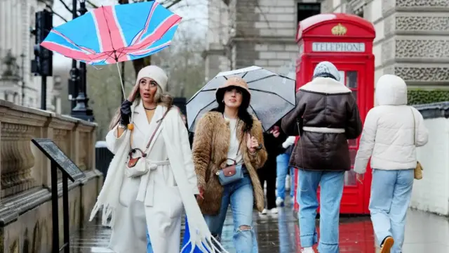 People hold umbrellas with the London skyline in front of them on a grey, rainy day