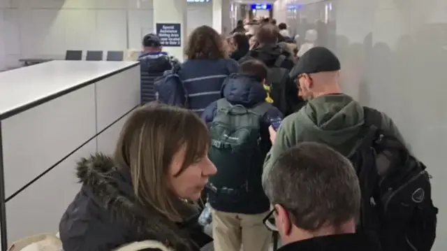 A line of people in coats and hats with bags inside a terminal at Bristol Airport