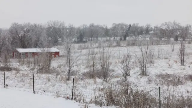 A red barn can be seen in a snowy farm field in Kansas. There is beige-coloured brush in the foreground and skies are grey.