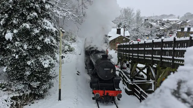 A steam train comes down the track with snow covering the ground. There are trees to the left and a fence to the right