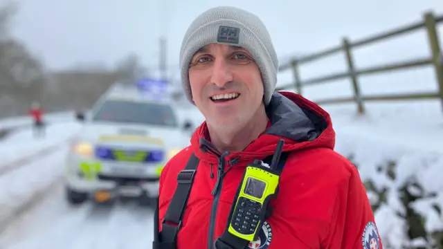 A smiling man wearing a grey hat and red jacket with a yellow emergency phone