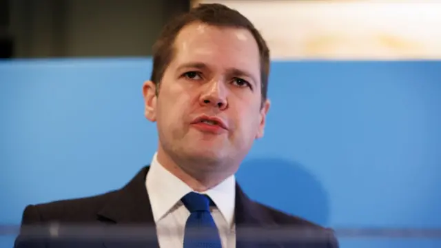 Robert Jenrick close up in black suit, blue tie and white shirt in front of a blue poster during the leadership campaign last autumn