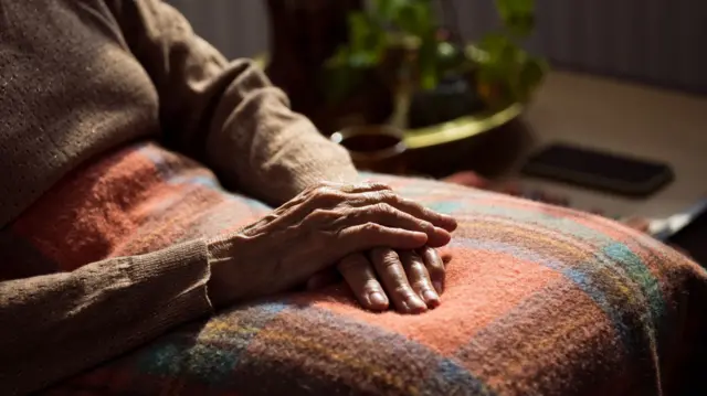 An elderly person sitting in a chair with a tartan orange, purple, blue and rust coloured blanket on their knee. The person's hands and torso are visible and they are wearing a beige jumper.