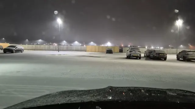 Picture taken from the front seat of a car at Bristol Airport. Snow is seen covering a nearly-empty car park, with bright floodlights illuminating the car park. In the distance, behind a wooden fence, bright lights of the runway are visible.