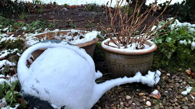 A watering can on its side and plant pots, both with a dusting of snow. A snowy field can be seen beyond.