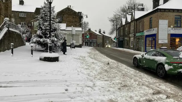 A car covered in snow drives through a gritted road in a town centre