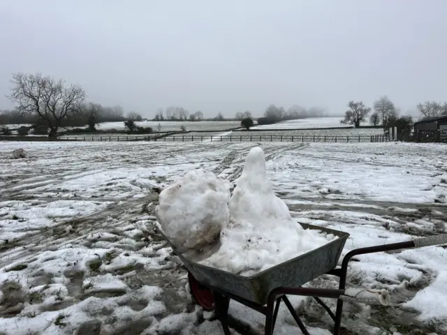 A snowy field in the Cotswolds, with a wheelbarrow in front of the camera loaded up with snow