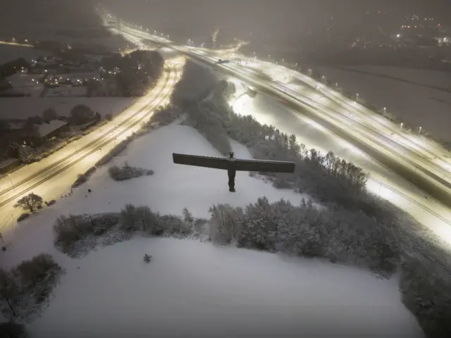 Aerial view of a large metal statue with large wings surrounded by snow and two well lit roads running either side