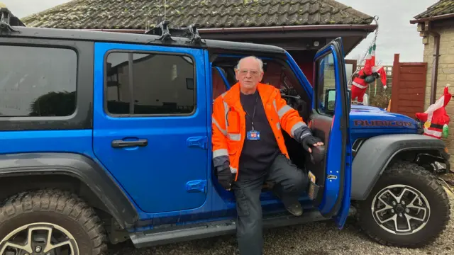 A wide view of Jerry, sat in the open door of his large, blue 4x4. He is wearing dark clothing, with an orange high-vis jacket over the top.