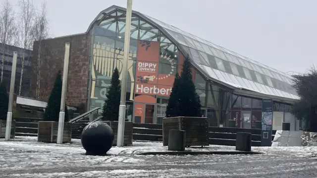 Snow thawing on a pedestrianised are in front of the Herbert art gallery and museum, which has a high arched glass roof and an advert for Dippy the dinosaur across its front window.