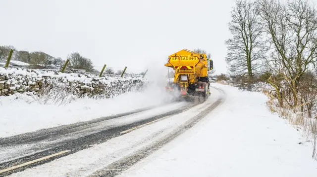 A yellow gritter drives through snow on a country road