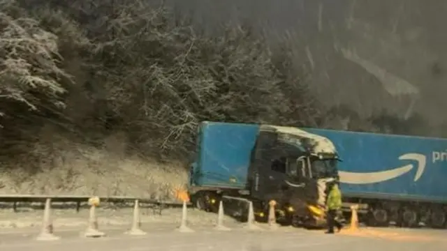 A jack knifed Amazon lorry on a motorway in the snow. A man in a yellow high-vis jacket stands beside it.