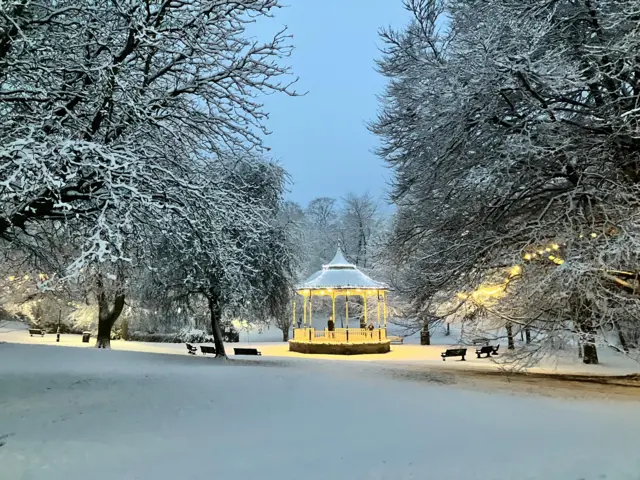 A bandstand lit by small lights around its top stand in a park surrounded by snow and large snow-covered trees