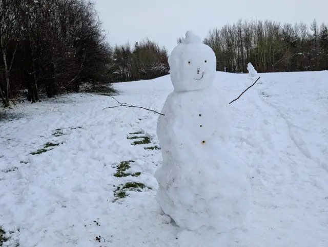 A large snowman in a field. He has twigs for arms