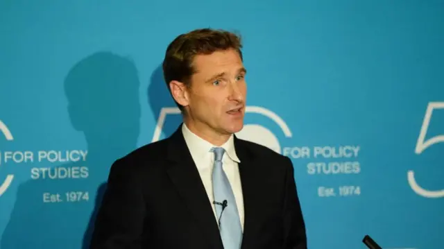 Chris Philp delivers speech standing in front of lectern wearing black suit, white shirt and light blue tie