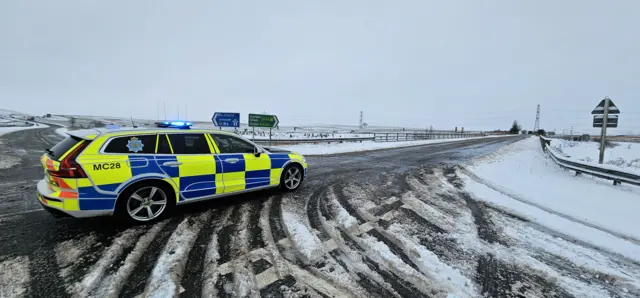 A Cumbria Police car on a snow-covered road junction. The care is in front of roadsign pointing to the M6 and to the A6