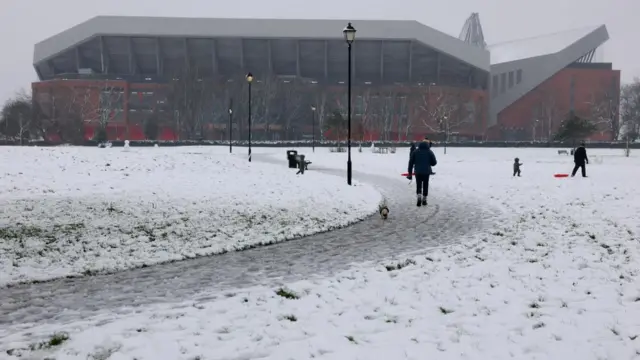 Snow on ground as people walk towards Anfield stadium. A man can be seen playing with two young children as he pulls a red sledge