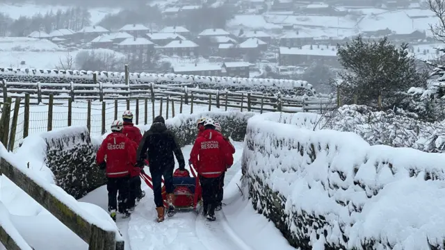 Five mountain rescue team members in red jackets and one in a black jacket walk along a snowy lane pushing a slegde with a person on it.