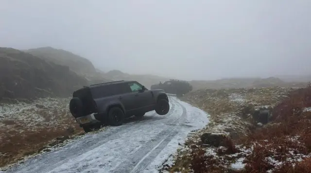 An icy single track road with two black cars run off it. The road is bordered by tall mountains.