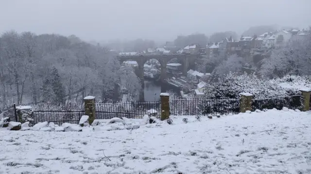 A snowy scene overlooking a river with a bridge. Trees to the left and houses to the right