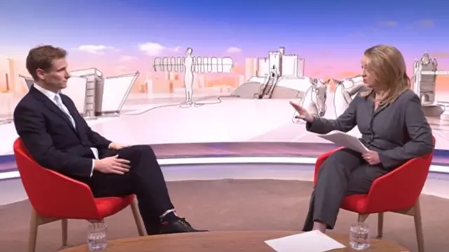 Chris Philp (L) sits on a red chair in a black suit while listening to Laura Kuenssberg (R) ask him questions. Kuenssberg is sitting of a red chair in a grey suit, a wooden table visible at the bottom of the frame with two glasses and sheets of paper on top