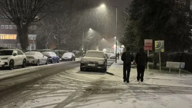 Two men in black walk away from camera, crossing road covered in snow with cars lining the street