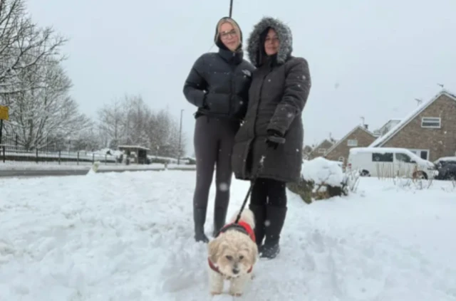 Two women pose for photo side by side over a pile of fresh snow, a small beige dog on a leash and warm covering in front of them. A white van covered in snow is visible behind the woman on the left, who has her winter jacket's hood pulled up. The other woman (L) has a black hoodie pulled up, strands of her blonde hair visible